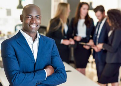 Black businessman leader looking at camera in modern office with multi-ethnic businesspeople working at the background. Teamwork concept. African man wearing blue suit.